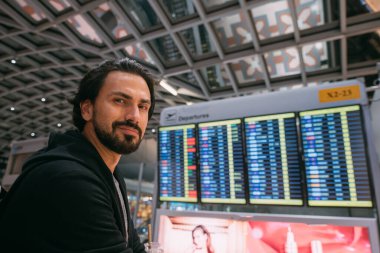 Havaalanındaki çizelgenin yanında bir adam var. Sırt çantalı genç bir adam terminaldeki uçuş programına bakıyor.