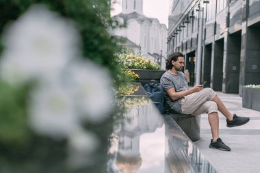 Bir adam büyük bir şehrin caddesinde bir telefonla bankta oturuyor. Genç beyaz yakışıklı adam metropolde yürüyerek yürüyor, yeşil bir şehir caddesinde akıllı bir telefonla dinleniyor ve iletişim kuruyor.