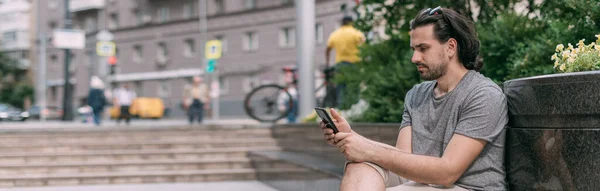 A man sits on a bench with a phone on the street of a big city. Young white handsome guy walks on foot in the metropolis, rests and communicates on a smartphone on a green city street