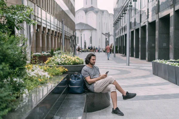 A man sits on a bench with a phone on the street of a big city. Young white handsome guy walks on foot in the metropolis, rests and communicates on a smartphone on a green city street