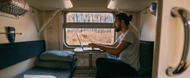 Trende dizüstü bilgisayarı olan bir adam. Portre. Genç yakışıklı adam tren yolunda uzaktan çalışıyor. Pencerenin yanındaki kompartımanda tek başına.