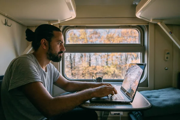 Man with laptop on the train. Portrait. Young handsome guy works remotely on a railway journey alone in a train compartment by the window