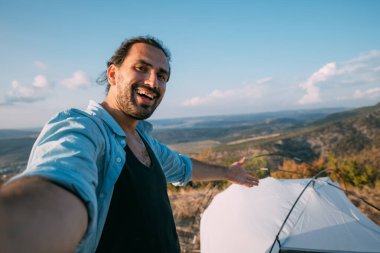 Genç erkek turist dağlardaki çadırın yakınında selfie çekiyor. Adam yürüyüş için kamp kurmuş, çadır kurmuş ve gün batımında dağlarda tek başına fotoğraf çekmiş.