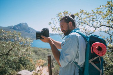 Bir adam, sırt çantalı ve kameralı bir turist dağlarda bir manzarayı filme alıyor. Kamp malzemesi ve kamera taşıyan genç adam yüksek kayalıkların ve mavi gökyüzünün yakınında fotoğraf ya da video çekiyor.