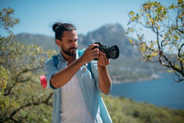 Erkek turist, deniz manzaralı bir dağ korusunda fotoğrafçı. Turist sırt çantalı ve kameralı genç yakışıklı ağaçlara bakıyor ve okyanusun manzarasına bakıyor.