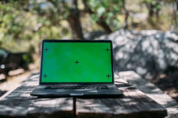 Open laptop on a wooden table in a camping in the mountains. A laptop with a green screen stands on a camping table outdoors among the trees against the background of a car with travel things