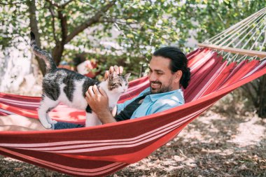 Genç bir adam bahçedeki bir hamakta dinleniyor ve bir kediyi okşuyor. Yakışıklı bir adam, güneşli bir yaz gününde, ağaçların gölgesinde bir kediyle bir hamakta uzanıyor.