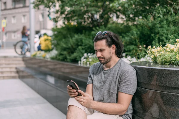男が大きな街の通りに電話をかけベンチに腰を下ろしている 若い白いハンサムな男は 大都市で徒歩で歩き 休憩し 緑の街の通りにスマートフォンで通信します — ストック写真