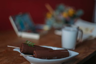 Suluboyalı, paletli, eskiz defterli bir ressam masası. Durgun hayat. Bahar çiçekli ahşap masa, boya malzemeleri, süt kupası ve dondurma.