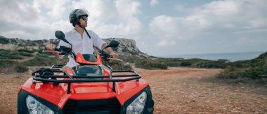 Bir adam kasklı ve dağlarda gözlüklü bir avluda oturuyor. Beyaz elbiseli yakışıklı bir adam ve kayaların arkasında kumlu bir yolda çeyrek bisiklet.