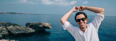 Denize bakan kayalıklarda bir adam. Beyaz elbiseli yakışıklı adam kayalık sahildeki okyanus manzarasının tadını çıkarıyor. # Denizin ötesindeki genç adam #