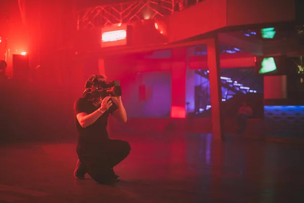 Director of photography with a camera in his hands on the set. Professional videographer at work on filming a movie, commercial or TV series. The filming process indoors, on a concert stage with neon light.