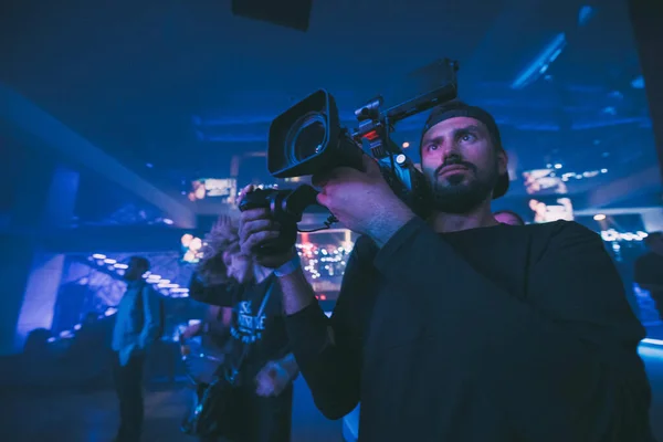 Director of photography with a camera in his hands on the set. Professional videographer at work on filming a movie, commercial or TV series. The filming process indoors, on a concert stage with neon light.