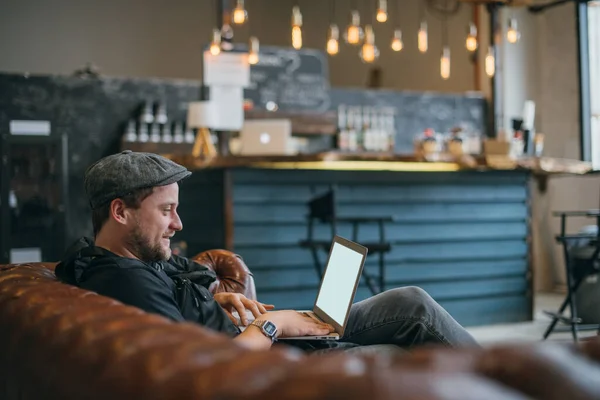 一个拿着笔记本电脑的男人在沙发上工作 一个年轻时髦的家伙一个人坐在电脑前 在室内的一个协作空间里远程工作 后续行动 — 图库照片