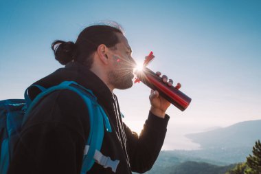 Yürüyüş yapan bir adam şafak vakti bir dağın tepesinde termostan çay içer. Turist ekipmanlı genç adam neşe içinde sabah güneşiyle denize bakan dağlarda buluşuyor.