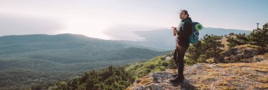 Yürüyüş yapan bir adam şafak vakti bir dağın tepesinde termostan çay içer. Turist ekipmanlı genç adam neşe içinde sabah güneşiyle denize bakan dağlarda buluşuyor.