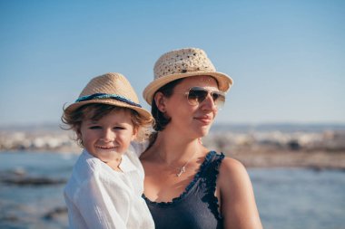 Deniz kenarında geziye çıkmış mutlu bir anne ve küçük bir oğlun portresi. Hasır şapkalı ve güneş gözlüklü anne kollarında şık bir başlık giymiş küçük bir çocuk tutuyor. Yakın plan..