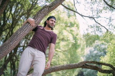 Parkta bir ağaçta genç bir adam oturuyor. Yaz kıyafetleri ve çıplak ayaklı yakışıklı adam yeşil bir parkta dinleniyor. Büyük bir söğüt ağacının üzerinde, yaz gününün tadını çıkarıyor.