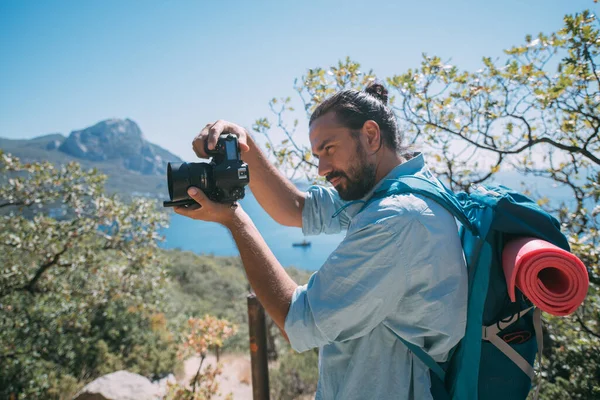 Bir adam, sırt çantalı ve kameralı bir turist dağlarda bir manzarayı filme alıyor. Kamp malzemesi ve kamera taşıyan genç adam yüksek kayalıkların ve mavi gökyüzünün yakınında fotoğraf ya da video çekiyor.