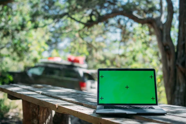 Open laptop on a wooden table in a camping in the mountains. A laptop with a green screen stands on a camping table outdoors among the trees against the background of a car with travel things