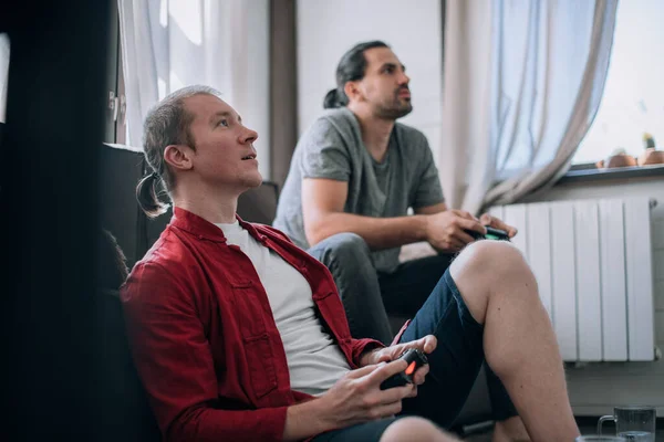 The guys are playing video games on the couch. Two young men with joysticks in their hands are enthusiastically playing a game on a console