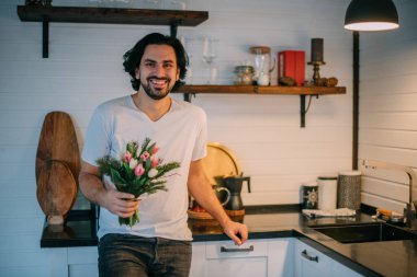 Mutfakta buketi olan yakışıklı bir adam. Uluslararası Kadınlar Günü. Genç bir adam 8 Mart 'ta elinde bir buket lale tutuyor. Evde romantik bir aile akşamı..