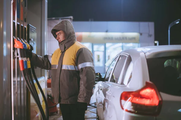 A worker at a gas station refueling a car. Young male refueller in uniform refueled a passenger car at a gas station at night