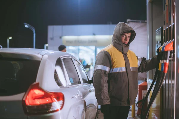 A worker at a gas station refueling a car. Young male refueller in uniform refueled a passenger car at a gas station at night