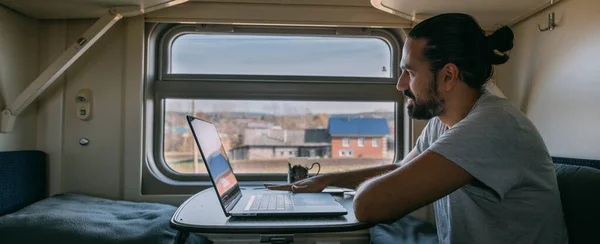 Man with laptop on the train. Portrait. Young handsome guy works remotely on a railway journey alone in a train compartment by the window