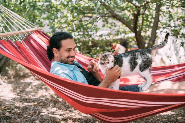 庭のハンモックに猫を撫でて休んでいる若い男 ハンサムな男は夏の晴れた日に木の木陰で猫とハンモックでリラックスしています — ストック写真