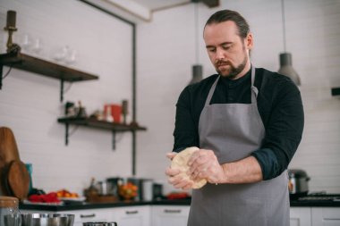 Erkek bir şef evde mutfakta hamur hazırlar. Mutfağındaki önlüklü genç adam. Masadaki hamur yapımında kullanılan malzemelerle birlikte.