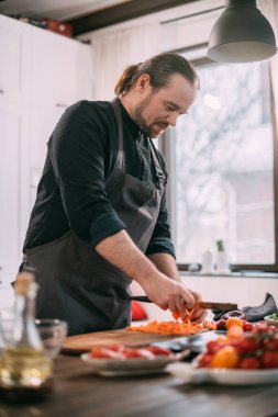 Erkek bir aşçı mutfakta evde sebze hazırlar. Evdeki mutfakta önlüklü genç adam sebze güveci ya da salata yapmak için malzemelerle birlikte.