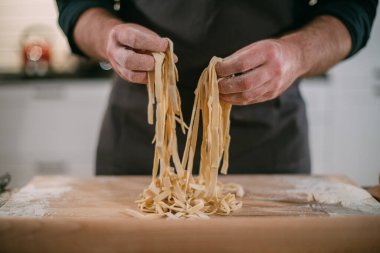 Erkek bir şef mutfakta erişte hazırlar. Merdaneli ve hamurlu eller. Evdeki mutfaktaki önlüklü genç adam. İçinde ev yapımı erişteler var.