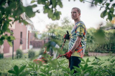 Bir adam bahçeyi hortumla suluyor. Parlak elbiseli, yeşil çiçekli bahçesinde su dolu bir hortumla çalışan genç adam.