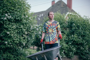 Bahçede çalışan bir adam, boş bir el arabasıyla yürüyor. Çiçekli çalıların arasında parlak iş elbiseleri ve eldivenleri olan genç bir adam.