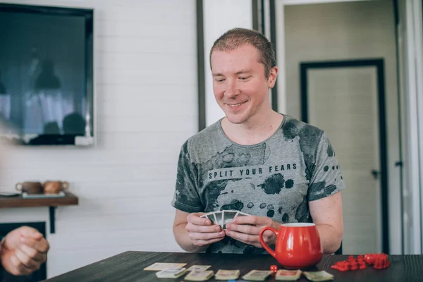 Friends play a board game in the living room. The company of young guys sits at a table and emotionally and cheerfully plays a card game on the weekend