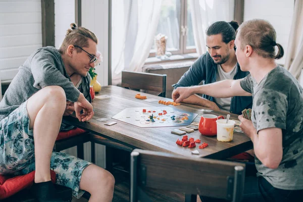 Friends play a board game in the living room. The company of young guys sits at a table and emotionally and cheerfully plays a card game on the weekend