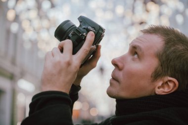 Elinde kamera olan bir adam kışın eski şehrin caddesini fotoğraflıyor. Arka planda sanatsal dekore edilmiş sokak lambalarının altında kamera ve eşarplı bir fotoğrafçı.