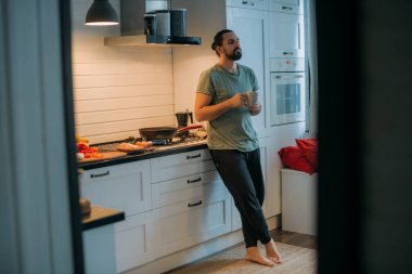 Bir adam mutfakta kahvaltı hazırlar. Genç beyaz erkek, büyük, parlak bir mutfakta, gaz sobasında öğle yemeği için yemek hazırlıyor.