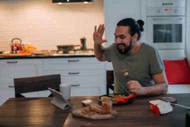 Bir adam evde tabletle kahvaltı yapıyor. Genç yakışıklı beyaz adam yemek yer, kahve içer, sabahın erken saatlerinde büyük, parlak bir mutfakta tablete bakar.