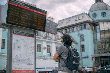 Bir adam istasyondaki tren saatlerine bakıyor. Sırt çantalı genç adam caddedeki büyük istasyonun yanındaki programa bakıyor.