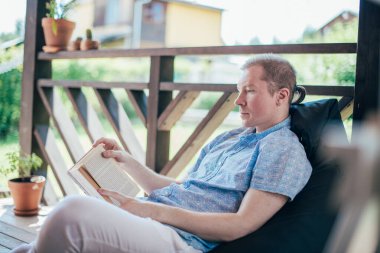 Bir adam verandada kitap okuyor. Genç yakışıklı bir adam güneşli bir günde elinde kitapla bir kır evinin terasında yatıyor.