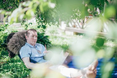 Bir adam hamakta dinleniyor ve kitap okuyor. İzin günü ve dinlenme. Genç yakışıklı bir adam, güneşli bir günde elinde kitapla bir hamakta, ağaçların gölgesinde yatıyor.