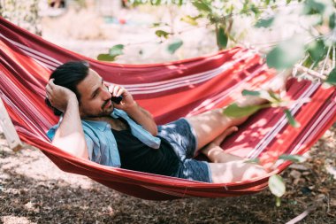 Genç bir adam dinleniyor ve bahçede bir hamakta telefonla konuşuyor. Yakışıklı adam bir hamakta uzanıyor ve bir yaz günü ağaçların gölgesinde akıllı bir telefonla iletişim kuruyor.