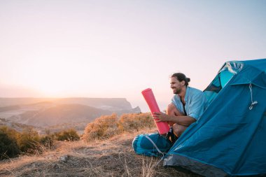 Dağlarda çadırda sırt çantalı genç bir erkek turist. Yürüyüş yaparken kampı terk eden bir adam, büyük bir sırt çantası ve güneş batarken dağlarda tek başına uyuyan bir köpükle tentenin altında oturuyor.