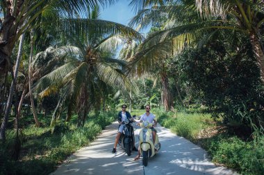 Tropik bir adada palmiye ağaçlarında motosiklet süren iki adam. Palmiye bahçesinde bisiklet süren turistler.