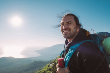 Gün doğumunda dağın tepesinde sırt çantalı bir erkek turist. Turist ekipmanlı genç adam neşe içinde sabah güneşiyle denize bakan dağlarda buluşuyor.