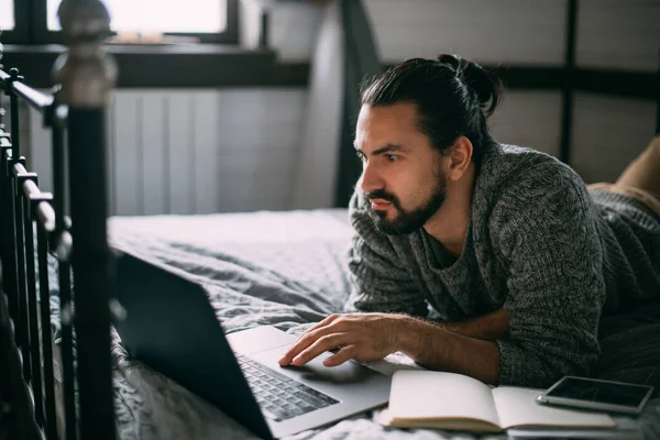 Young man lies on bed with laptop at home. Handsome, relaxed Caucasian man works, studies, surfs the net in the bedroom on a comfortable bed during the day