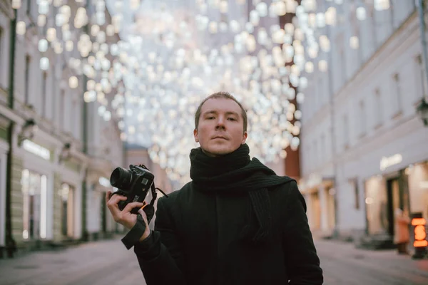 A man with a camera photographs the street of the old city in winter. Photographer with a camera in a coat and scarf on the background of artfully decorated street lamps