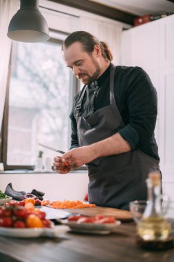 Erkek bir aşçı mutfakta evde sebze hazırlar. Evdeki mutfakta önlüklü genç adam sebze güveci ya da salata yapmak için malzemelerle birlikte.